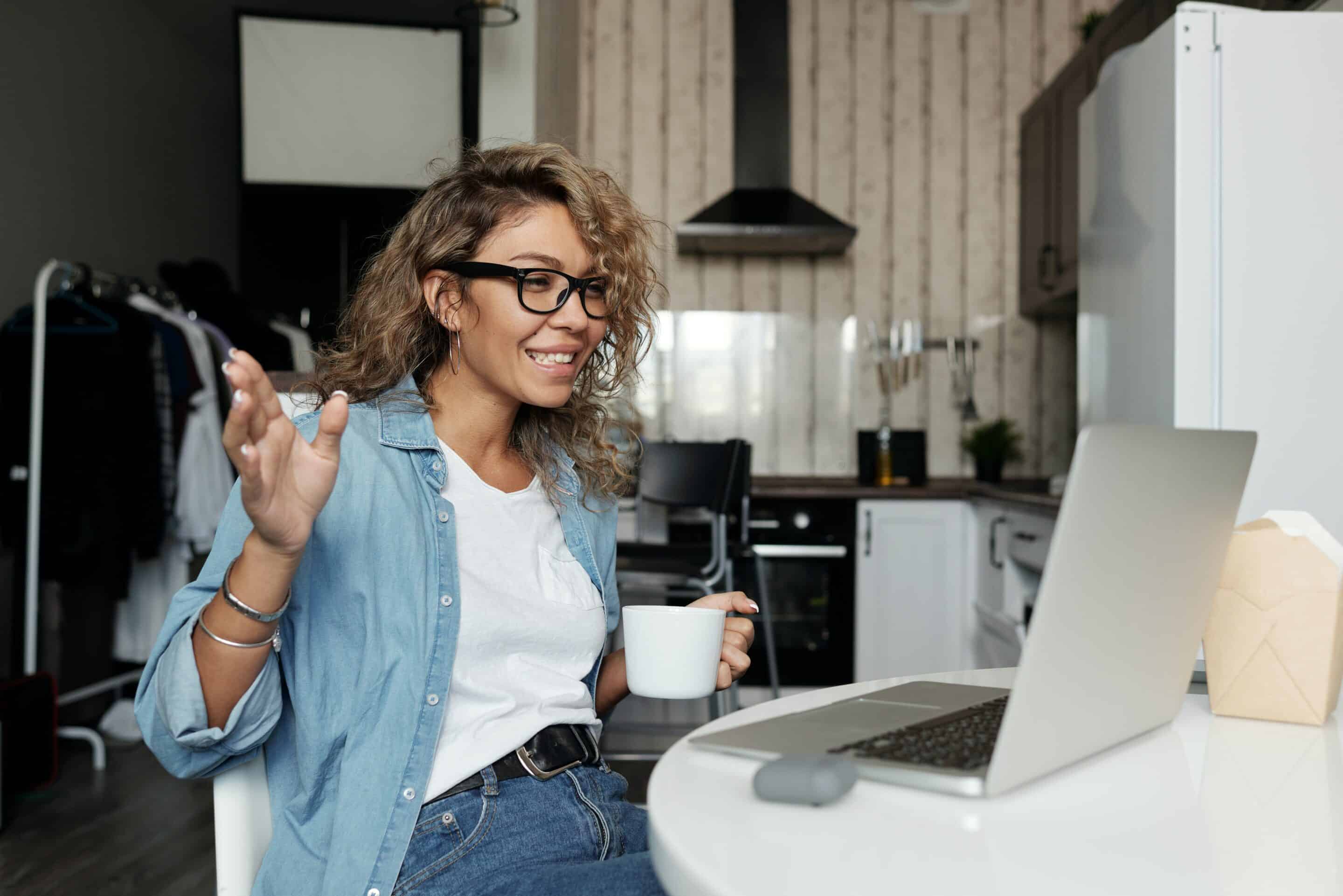 smiling girl with glasses and curly hair holding coffee cup sitting in front of computer, joining a virtual trauma processing group